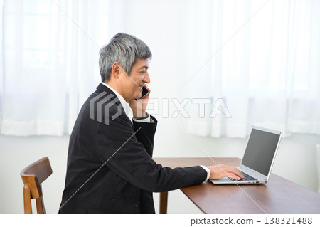 一位六十多歲的男子一邊用智慧型手機通話，一邊看著筆記型電腦。 138321488