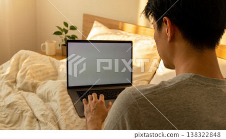 Candid documentary of a diverse young man working on a laptop in bed with natural light, capturing a real life morning routine, remote work lifestyle, and everyday work life balance at home 138322848