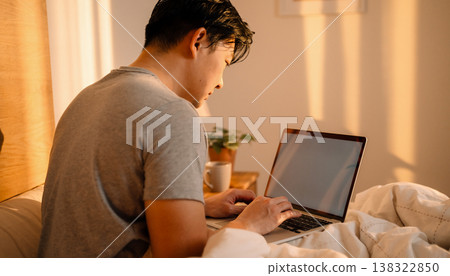 Candid documentary of a diverse young man working on a laptop in bed with natural light, capturing a real life morning routine, remote work lifestyle, and everyday work life balance at home 138322850