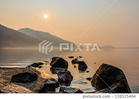 The dawn over Lake Shikotsu, bathed in orange light; a tranquil lakescape guided by stones. The dawn over Lake Shikotsu, bathed in orange light; a tranquil lakescape guided by stones. 138325404