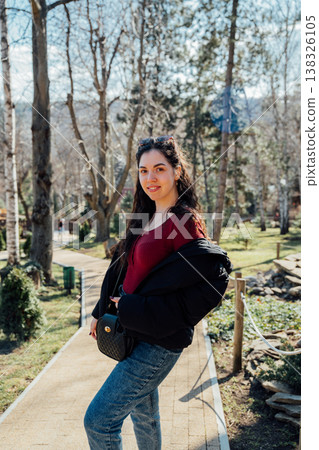 Young woman of slavic appearance posing on park pathway wearing red top jeans and black jacket holding handbag in sunny outdoor natural setting Young woman of slavic appearance posing on park pathway wearing red top jeans and black jacket holding handbag in sunny outdoor natural setting 138326105