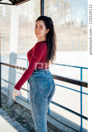 Full body portrait of smiling slavic woman with long curly dark hair wearing red top and blue jeans posing near railing in urban outdoor setting 138326112