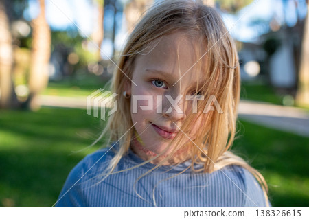 Young girl with blonde hair and blue eyes smiles playfully outdoors in a park, surrounded by green grass and blurred trees in the background Young girl with blonde hair and blue eyes smiles playfully outdoors in a park, surrounded by green grass and blurred trees in the background 138326615