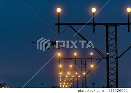 Airplane approaching landing with illuminated runway lights against a twilight sky, showcasing the airport's approach lighting system and surrounding infrastructure Airplane approaching landing with illuminated runway lights against a twilight sky, showcasing the airport's approach lighting system and surrounding infrastructure 138326631