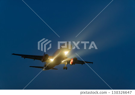 Airplane flying at night with landing lights illuminated, showcasing the aircraft's silhouette against a dark blue sky with faint clouds and atmospheric lighting effects 138326647