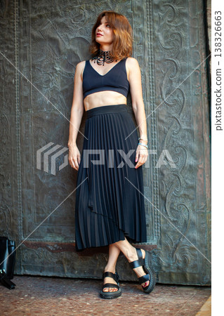 Female model wearing black crop top and pleated skirt poses against an ornate textured wall, showcasing a contemporary fashion style with sandals and accessories 138326663