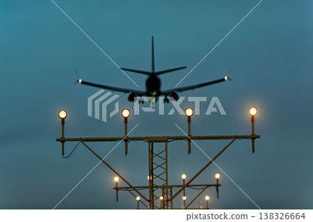 Airplane approaching landing with illuminated runway lights visible in the foreground against a cloudy twilight sky creating a dramatic aviation scene Airplane approaching landing with illuminated runway lights visible in the foreground against a cloudy twilight sky creating a dramatic aviation scene 138326664