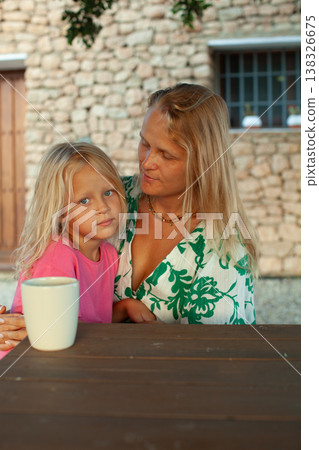 Caucasian woman and girl sitting at wooden table outdoors, with stone wall and window in the background, enjoying a moment together with a white cup Caucasian woman and girl sitting at wooden table outdoors, with stone wall and window in the background, enjoying a moment together with a white cup 138326675
