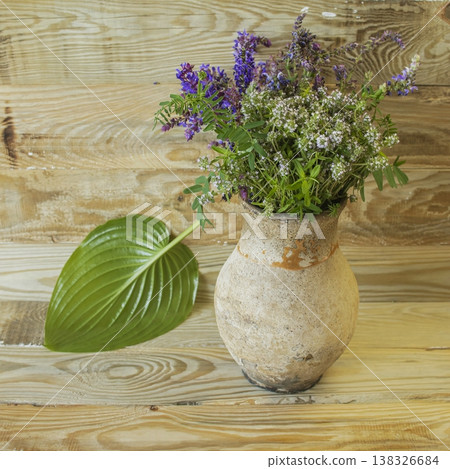 Rustic still life with wildflowers in a clay vase and a hosta leaf on a wooden background. Rustic still life with wildflowers in a clay vase and a hosta leaf on a wooden background. 138326684