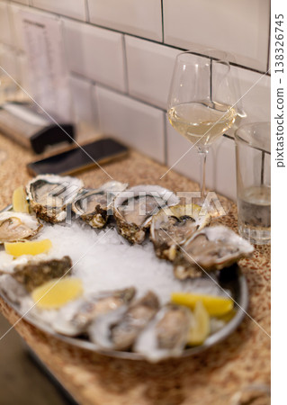 Fresh oysters served on ice with lemon wedges alongside glasses of white wine in a modern restaurant setting with tiled walls and wooden counter 138326745
