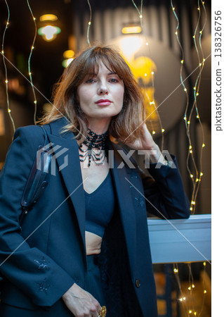 Stylish woman with brown hair wearing black outfit and choker poses near window adorned with decorative lights in a modern urban setting Stylish woman with brown hair wearing black outfit and choker poses near window adorned with decorative lights in a modern urban setting 138326756