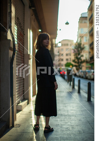Young woman with shoulder-length hair wearing black outfit stands on urban street during sunset, buildings and parked cars visible in the background 138326762
