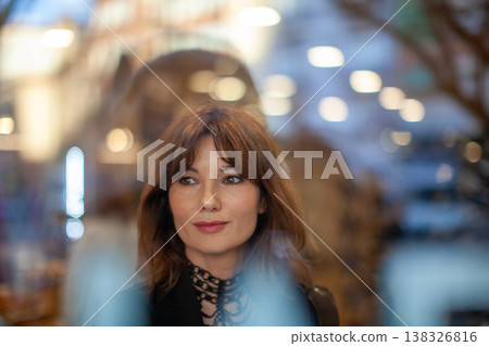 Female with brown hair and light makeup gazes thoughtfully through a glass window, with blurred lights and urban scenery visible in the background Female with brown hair and light makeup gazes thoughtfully through a glass window, with blurred lights and urban scenery visible in the background 138326816