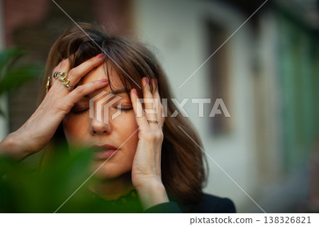 Woman with brown hair wearing rings holds her head with both hands, eyes closed, surrounded by greenery and blurred buildings in the background Woman with brown hair wearing rings holds her head with both hands, eyes closed, surrounded by greenery and blurred buildings in the background 138326821