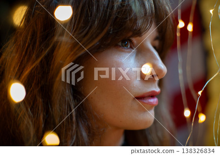 Female model with long brown hair and natural makeup poses against a backdrop of warm fairy lights, creating a soft and inviting atmosphere in an indoor setting Female model with long brown hair and natural makeup poses against a backdrop of warm fairy lights, creating a soft and inviting atmosphere in an indoor setting 138326834