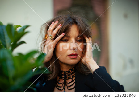 Woman with dark hair wearing black attire and jewelry poses with hands on her head, surrounded by green foliage in an outdoor setting with soft natural lighting 138326846