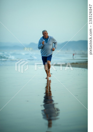 Older man jogging barefoot along a sandy beach shoreline, wearing a light blue sweatshirt and shorts, with ocean waves and distant figures in the background 138326847