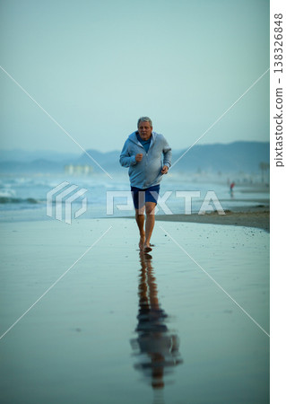 Senior man jogging along the beach shoreline at sunrise, wearing a light blue hoodie and shorts, with reflections visible in the wet sand and waves in the background 138326848