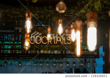 Neon sign reading "COCKTAILS" illuminated above a bar with hanging light bulbs and shelves stocked with various bottles and glassware in a modern setting 138326851