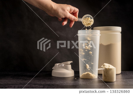 Protein drink. Hand pouring protein powder in a white shaker. Plastic tube on dark background. Body building, muscle growth and food supplement concept Protein drink. Hand pouring protein powder in a white shaker. Plastic tube on dark background. Body building, muscle growth and food supplement concept 138326860