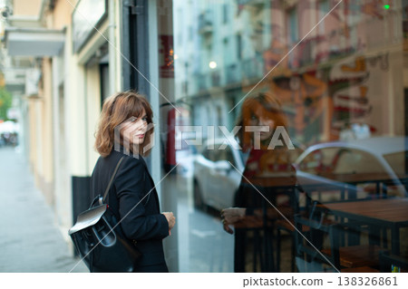 Female figure with brown hair wearing black attire stands outside a cafe, gazing at her reflection in the glass window, urban street scene visible in the background Female figure with brown hair wearing black attire stands outside a cafe, gazing at her reflection in the glass window, urban street scene visible in the background 138326861