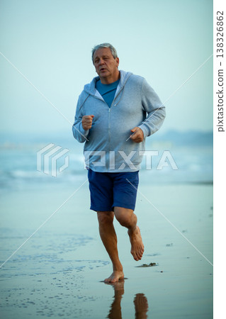 Senior man jogging barefoot along the shoreline in a gray hoodie and blue shorts, with gentle waves and a hazy beach background during early morning light Senior man jogging barefoot along the shoreline in a gray hoodie and blue shorts, with gentle waves and a hazy beach background during early morning light 138326862