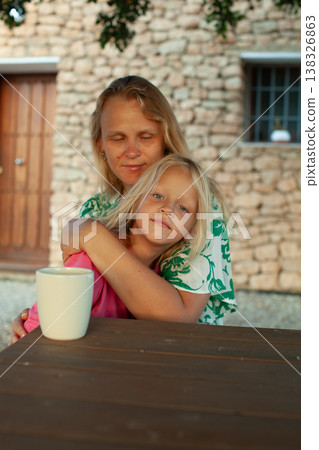 Caucasian woman and child embrace at a wooden table with a white mug, stone wall and wooden door visible in the background, outdoor setting with natural light Caucasian woman and child embrace at a wooden table with a white mug, stone wall and wooden door visible in the background, outdoor setting with natural light 138326863