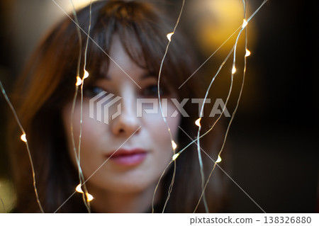 Woman with brown hair gazes softly through glowing string lights, creating a warm ambiance in a dimly lit indoor setting with blurred background elements Woman with brown hair gazes softly through glowing string lights, creating a warm ambiance in a dimly lit indoor setting with blurred background elements 138326880