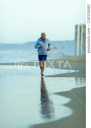 Senior man jogging barefoot along the shoreline at dawn, with ocean waves gently lapping at his feet and tall buildings visible in the background 138326882