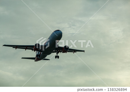 Commercial airplane flying through cloudy sky during sunset, showcasing landing gear extended and wings positioned for descent against a dramatic backdrop 138326894