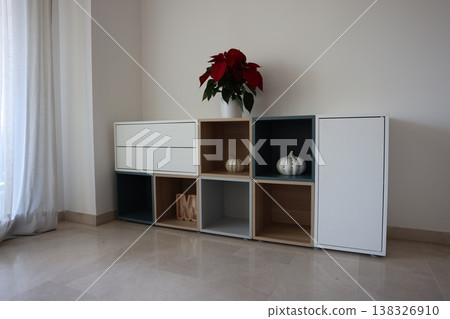 Modern furniture unit with white and wooden compartments, featuring decorative items like a pumpkin and a poinsettia plant in a bright interior space Modern furniture unit with white and wooden compartments, featuring decorative items like a pumpkin and a poinsettia plant in a bright interior space 138326910