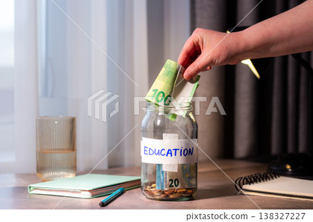 Hand dropping money into glass savings jar on table. Close up of fingers inserting cash into transparent container for education. Financial planning. University. 138327227