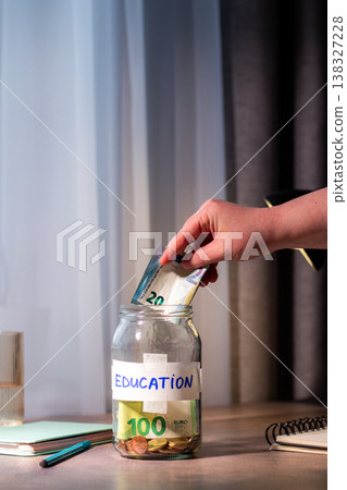 Hand dropping money into glass savings jar on table. Close up of fingers inserting cash into transparent container for education. Financial planning. University. Vertical 138327228