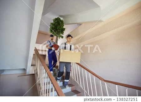 Two professional movers in uniform carrying box and potted plant downstairs in apartment building. 138327251