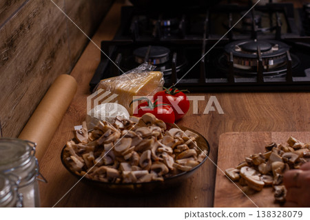 Fresh chopped mushrooms, whole tomatoes, and cheese blocks are ready on a wooden kitchen counter next to a gas stove, featuring a hand using a knife and a cutting board. 138328079
