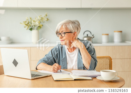 Middle aged senior woman using laptop computer writing notes at home. Focused mature old businesswoman work in home office making written records doing online work. Mature boss professional worker 138329163