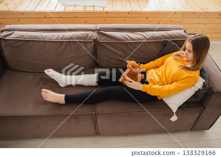 Young girl with broken leg cast sitting on sofa. Sad little kid hugging her teddy bear recovering from broken leg at home. Girl using cast and crutches. Accident injury fracture treatment foot bandage Young girl with broken leg cast sitting on sofa. Sad little kid hugging her teddy bear recovering from broken leg at home. Girl using cast and crutches. Accident injury fracture treatment foot bandage 138329166