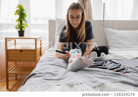 Young teenage girl with broken leg sitting on bed draws with felt tip pens on orthopedic cast. Bored girl with broken leg playing at home during rehabilitation. Accident injury fracture treatment Young teenage girl with broken leg sitting on bed draws with felt tip pens on orthopedic cast. Bored girl with broken leg playing at home during rehabilitation. Accident injury fracture treatment 138329174