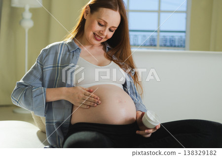 Pregnancy motherhood. Pregnant woman applying cream lotion on big belly takes care of her skin. Cosmetic product for prevention stretch mark. Girl enjoying pregnancy at home. Maternity health concept 138329182