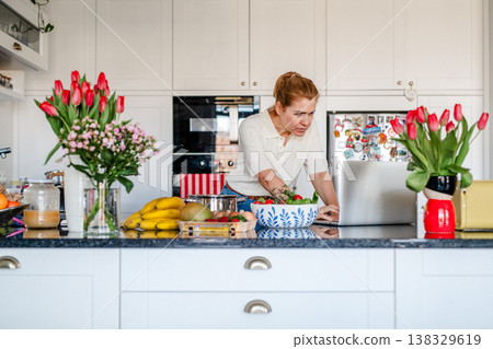 Mature woman searching recipe online on laptop while cooking in kitchen. Mature woman searching recipe online on laptop while cooking in kitchen. 138329619
