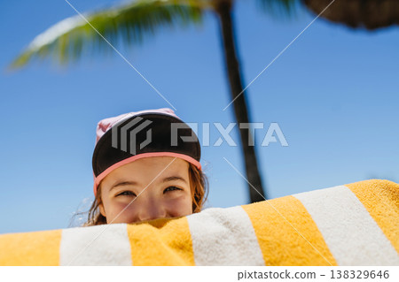 Smiling child peeking over beach towel under palm tree. Smiling child peeking over beach towel under palm tree. 138329646