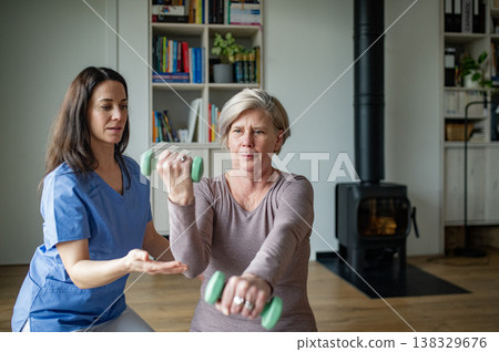 Physiotherapist assisting senior woman with dumbbell exercise, home rehabilitation. Physiotherapist assisting senior woman with dumbbell exercise, home rehabilitation. 138329676