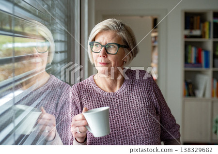 Elderly woman having cup of tea and looking through window at home. 138329682
