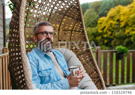 Man is enjoying cup of warm tea, coffee, having relaxing moment at home, sitting in hanging chair on patio. Man is enjoying cup of warm tea, coffee, having relaxing moment at home, sitting in hanging chair on patio. 138329746