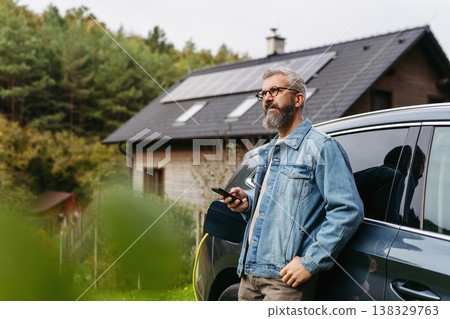 Charging electric car at home, using app. Solar panels on roof behind him. Charging electric car at home, using app. Solar panels on roof behind him. 138329763