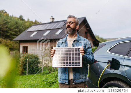 Man holding model of solar panel in hands. Charging electric car and house with solar panel system on roof behind him. 138329764