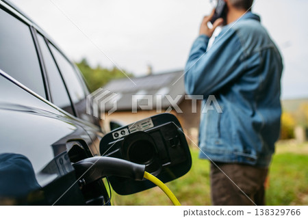 Man charging electric car in front of his house while making phone call. Man charging electric car in front of his house while making phone call. 138329766