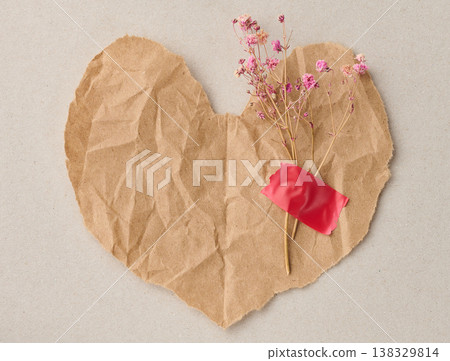 A torn piece of crumpled craft paper in the shape of a heart with a sprig of pink dried flowers glued with red tape A torn piece of crumpled craft paper in the shape of a heart with a sprig of pink dried flowers glued with red tape 138329814