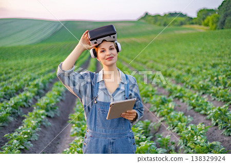 A woman in overalls and a plaid shirt stands in a field of crops, wearing a VR headset and holding a tablet, symbolizing modern agricultural technology A woman in overalls and a plaid shirt stands in a field of crops, wearing a VR headset and holding a tablet, symbolizing modern agricultural technology 138329924