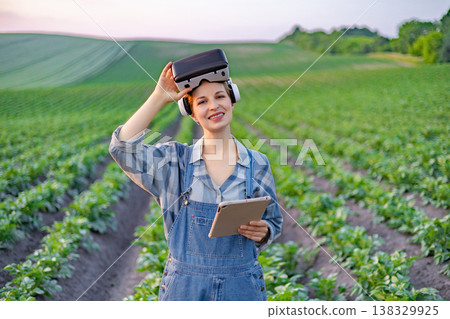 A woman in overalls and a plaid shirt stands in a field of crops, wearing a VR headset and holding a tablet, symbolizing modern agricultural technology A woman in overalls and a plaid shirt stands in a field of crops, wearing a VR headset and holding a tablet, symbolizing modern agricultural technology 138329925